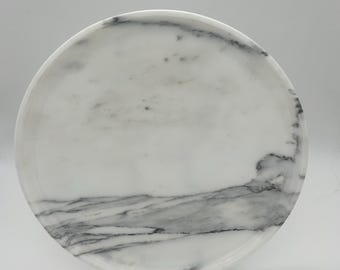 May include: A round white marble serving tray with gray veining. The tray is on a black stand.