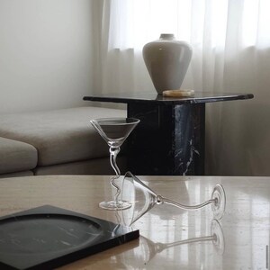 May include: Two clear glass martini glasses with a curved stem on a light beige table with a black marble tray. The glasses are empty and are sitting on a table with a white vase and a black marble side table in the background.