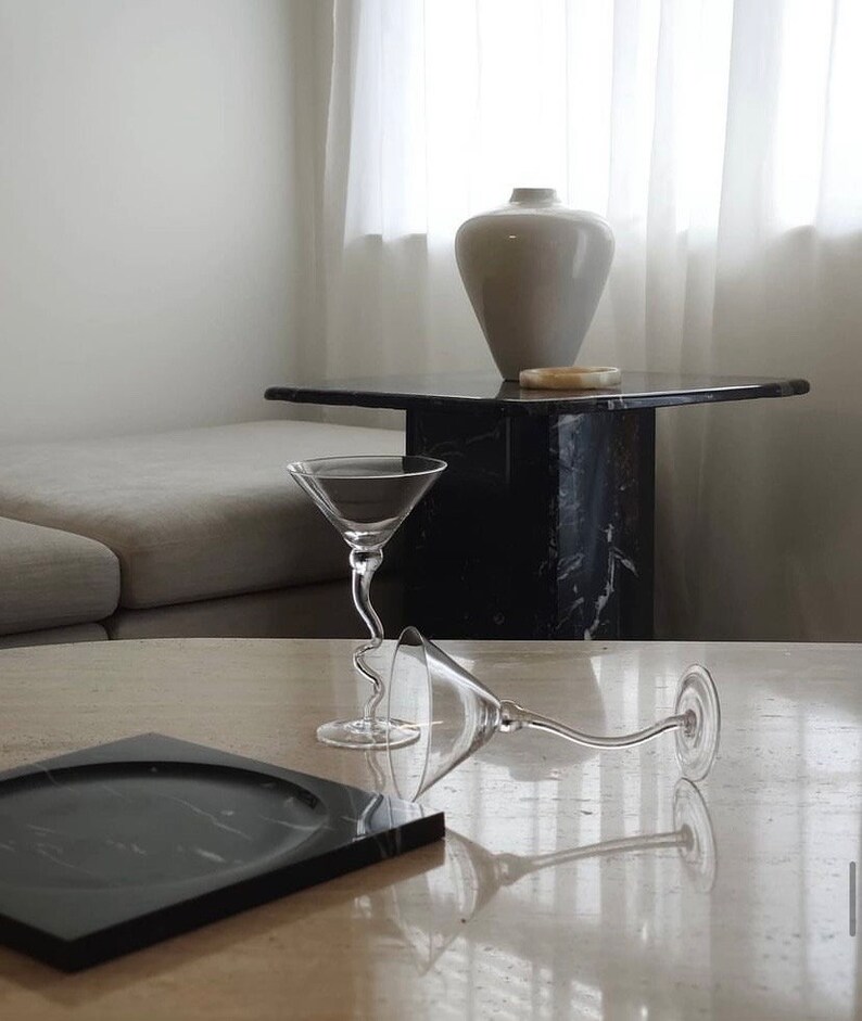 May include: Two clear glass martini glasses with a curved stem on a light beige table with a black marble tray. The glasses are empty and are sitting on a table with a white vase and a black marble side table in the background.