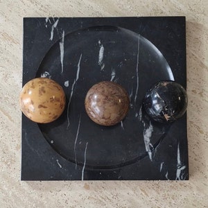 May include: A black marble tray with a rounded edge and three decorative spheres. The spheres are made of different types of marble, one is brown and white, one is black and white, and one is tan and brown.