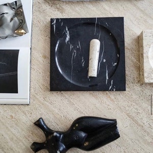 May include: A black marble mortar and pestle set with a white marble pestle. The mortar has a round, slightly indented surface. The set is on a light beige surface.