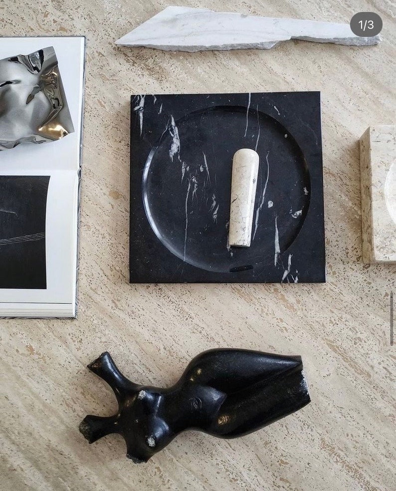 May include: A black marble mortar and pestle set with a white marble pestle. The mortar has a round, slightly indented surface. The set is on a light beige surface.