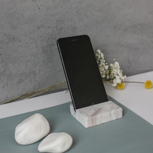 Pode incluir: Um smartphone preto está apoiado em um suporte de telefone de mármore branco. O suporte está sobre uma superfície azul esverdeada com duas pedras brancas.