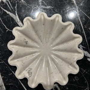 May include: A light gray, stone-like, decorative dish with a scalloped edge. The dish has a sunburst design with radiating lines from the center. The dish is on a black and white marble surface.