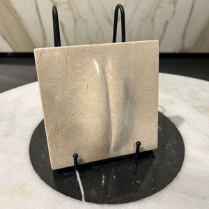 May include: A beige square stone tile with a curved, indented design, displayed on a black metal stand. The stand is on a black circular base, set against a marble surface. The tile's texture is visible.