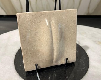 May include: A beige square stone tile with a curved, indented design, displayed on a black metal stand. The stand is on a black circular base, set against a marble surface. The tile's texture is visible.