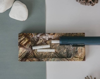May include: A rectangular ashtray with a natural stone pattern holds three cigarettes and a dark teal electronic cigarette. Two white stones and a pine cone are in the background.