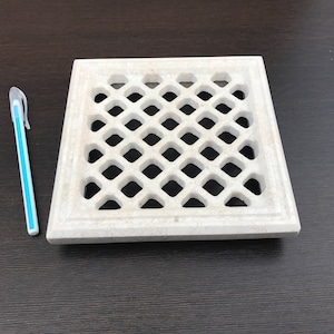 May include: A white square vent cover with a diamond-shaped grid pattern. The vent cover is made of a textured material and has a slightly raised border.