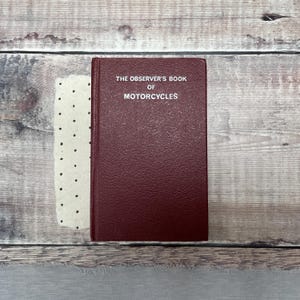 May include: A maroon hardback book titled The Observer's Book of Motorcycles with a textured cover, positioned next to a cream-coloured paper insert featuring a repeating pattern of small black dots.