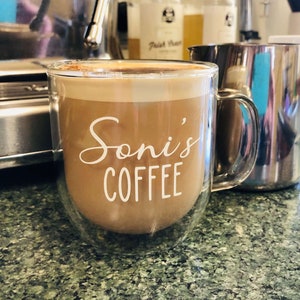 May include: A double-walled glass mug with a handle, filled with coffee and a layer of foam on top. The mug has the text "Soni's Coffee" printed on the side in white.