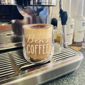 A clear, double walled coffee mug on a coffee machine. The mug is personalised with the words Ben's Coffee and filled with coffee.