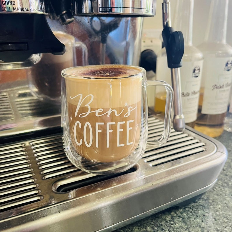 A clear, double walled coffee mug on a coffee machine. The mug is personalised with the words Ben's Coffee and filled with coffee.