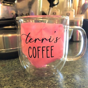 May include: A clear glass double-walled mug with a pink paper liner that says "Terri's Coffee" in black lettering.