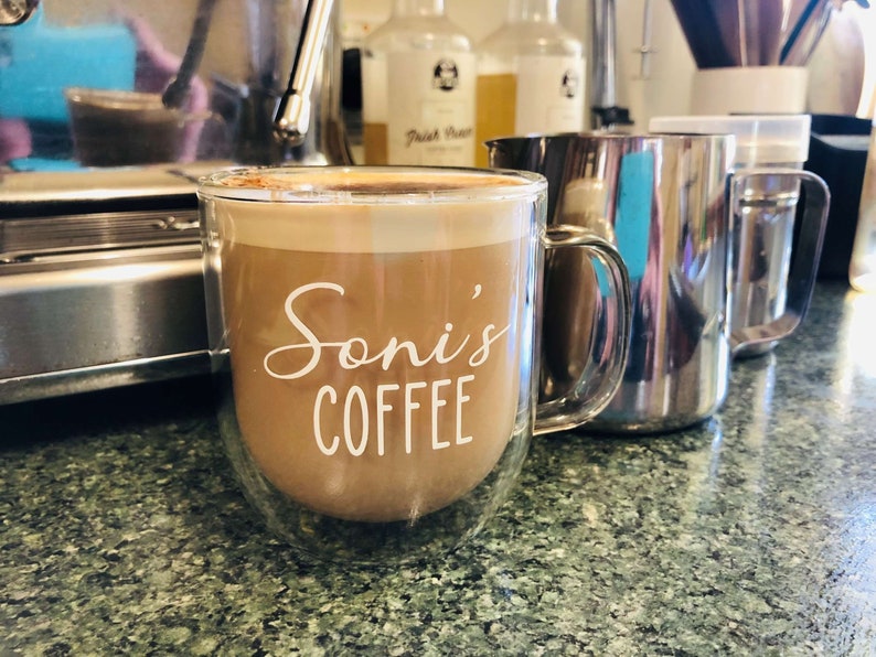 May include: A double-walled glass mug with a handle, filled with coffee and a layer of foam on top. The mug has the text "Soni's Coffee" printed on the side in white.
