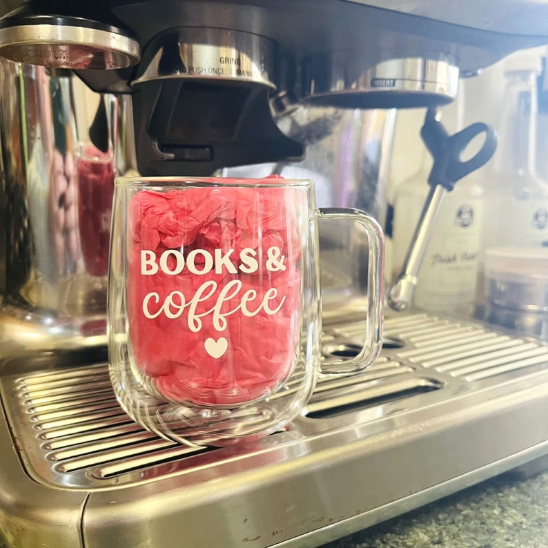 May include: A clear glass mug with a double wall design, featuring the text "BOOKS & coffee" in white lettering with a small heart symbol. The mug is sitting on a stainless steel surface.