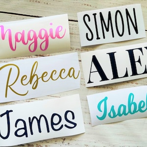 May include: Five white labels with different names written in various colours and fonts. The names are Maggie in pink, Rebecca in gold, Simon in black, Alex in black, and Isabella in turquoise.