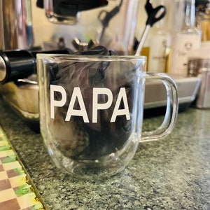 May include: A clear glass mug with a double wall design. The mug has the word "PAPA" printed in white on the front.