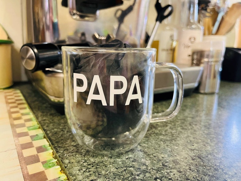 May include: A clear glass mug with a double wall design. The mug has the word "PAPA" printed in white on the front.