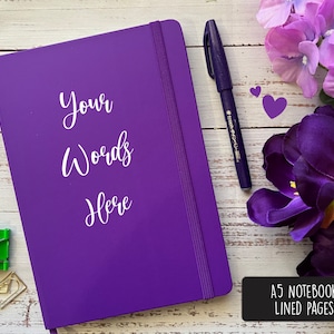 May include: Purple A5 notebook with white text that reads "Your Words Here" on the cover. The notebook is on a white wooden surface with a purple flower, a black pen, and two gold paper clips.