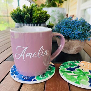 May include: A mauve ceramic mug with the name "Amelia" in white script, on a floral coaster. The coaster has blue and green floral patterns. Decorative plants in pots are in the background.
