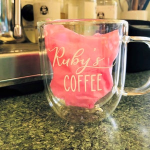 May include: A clear glass double-walled mug with a pink silicone sleeve that says "Ruby's Coffee" in white lettering.