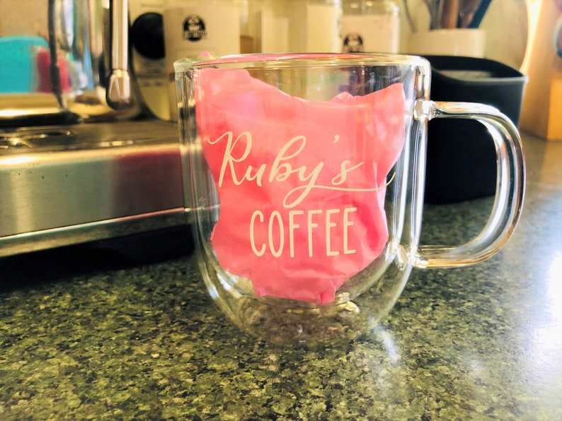 May include: A clear glass double-walled mug with a pink silicone sleeve that says "Ruby's Coffee" in white lettering.