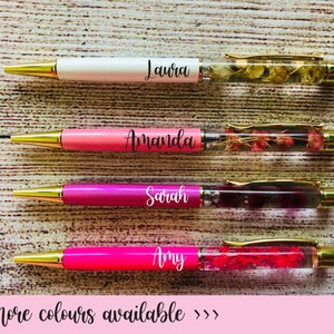 May include: Four personalised pens with different coloured bodies and clear barrels filled with dried flowers. The pens are labelled "Laura", "Amanda", "Sarah", and "Amy".
