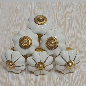 May include: Six white ceramic cabinet knobs with a flower design. Each knob has a gold-coloured centre and gold accents. The knobs are arranged in a pyramid shape on a light wood-grain patterned surface.