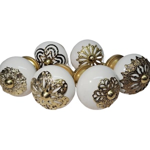 Op de afbeelding: Een verzameling witte keramische kastknoppen met goudkleurige metalen accenten. De knoppen hebben verschillende decoratieve ontwerpen, waaronder bloemen- en geometrische patronen. Beslag voor meubels en woondecoratie.