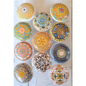 May include: A collection of twelve decorative ceramic cabinet knobs. Each knob features a unique, colorful, and intricate design with patterns in shades of orange, blue, yellow, and black. The knobs are round and have a glossy finish.