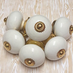 May include: A collection of white ceramic cabinet knobs with gold-colored metal accents. The knobs have a rounded shape and a decorative gold rosette design. The hardware is arranged on a light wood-grain patterned surface.