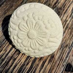 Könnte beinhalten: Ein weißer Keramik-Anhänger mit einem erhabenen Blumenmuster. Der Anhänger ist rund und hat eine strukturierte Oberfläche.