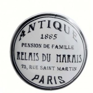 Könnte beinhalten: Weißer Keramik-Knopf mit schwarzem Rand und schwarzem Text, der "Antique 1885 Pension de Famille Relais du Marais 73, Rue Saint Martin Paris" lautet.