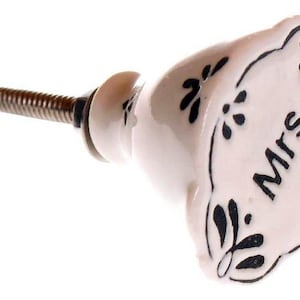 May include: White ceramic cabinet knob with black floral designs and the word "Mrs." written on it.