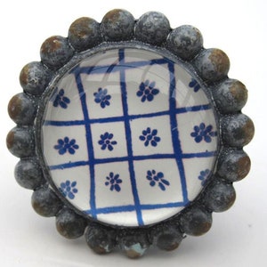 Könnte beinhalten: Runder, dekorativer Schrankknopf mit einem transparenten Glasmittelteil, das ein blaues Blumengittermuster aufweist. Der Knopf ist von einem dunklen, strukturierten Metallrand mit kleinen, abgerundeten Akzenten umgeben.