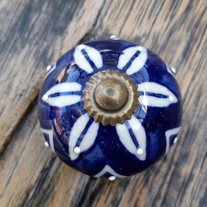May include: A round, blue ceramic cabinet knob with a white floral pattern. The knob has a brass-colored center and is set against a wooden surface. The design features a repeating pattern of white petal-like shapes.