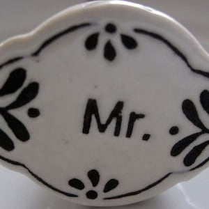 Può includere: Segnaposto ovale in ceramica bianca con disegni floreali neri e la parola "Mr." in lettere nere.