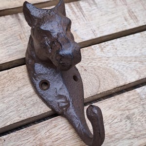 Pode incluir: Um gancho de parede de ferro fundido castanho escuro em forma de cabeça de cão. O gancho tem um acabamento texturizado e dois orifícios de montagem. A cabeça do cão é detalhada com orelhas, olhos e focinho. O gancho foi concebido para pendurar objetos.