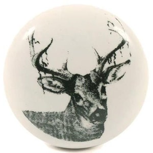 May include: A white, round cabinet knob featuring a detailed black and gray illustration of a deer's head with prominent antlers. The design is centered on the knob, creating a rustic and nature-inspired aesthetic. This decorative knob is suitable for furniture and home decor.