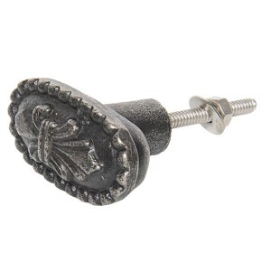 Könnte beinhalten: Ein silberfarbener Metall-Schrankknauf mit ovaler Basis und einem dekorativen Fleur-de-lis-Design. Der Knauf hat eine Schraube zum Befestigen an einer Schranktür.