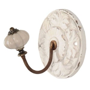Könnte beinhalten: Ein weißer Wandhaken mit floral gemusterter Rückplatte im Used-Look. Der Haken hat einen cremefarbenen, kürbisförmigen Knopf und einen gebogenen, bronzefarbenen Metallhaken. Das Design hat eine Vintage- und Antik-Ästhetik.
