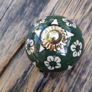 Pode incluir: Um puxador de armário decorativo e redondo. O puxador é verde escuro com desenhos florais brancos e um centro dourado. O puxador é provavelmente feito de cerâmica ou porcelana.