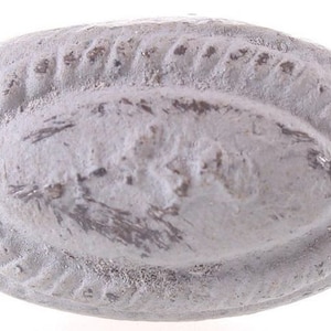 Könnte beinhalten: Ein weißer, ovaler Keramikknopf mit einem erhabenen, strukturierten Design am Rand. Die Mitte des Knopfes hat einen leicht vertieften Bereich mit einem verblassten, dunkelgrauen Design.