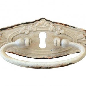 Pode incluir: Um puxador de gaveta oval de metal com alça pendente e fechadura central. A ferragem apresenta um acabamento esbranquiçado desgastado com bordas castanho-escuras e detalhes florais em relevo ao longo da borda superior da placa traseira.
