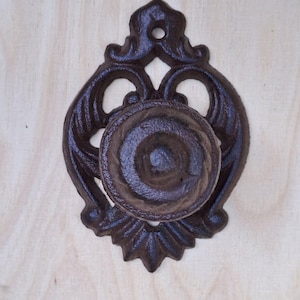 Puede incluir: Un pomo de puerta de hierro fundido de color marrón oscuro con un diseño decorativo de desplazamiento. El pomo está montado en una superficie de madera.
