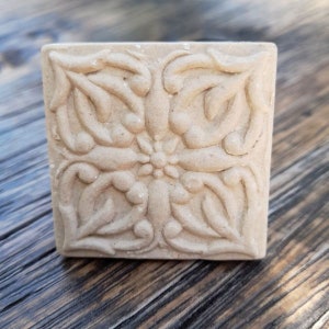 Könnte beinhalten: Eine quadratische, beigefarbene Dekorplatte mit einem erhabenen, filigranen Blumendesign. Die Platte hat eine strukturierte Oberfläche und eine leicht raue Oberfläche.