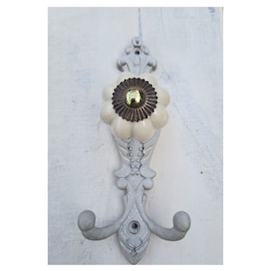 Op de afbeelding: Een zilveren metalen haak met een witte keramische knop met een gouden middelpunt. De haak heeft een decoratief bloemmotief en twee haken.