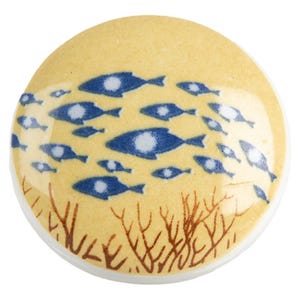 Op de afbeelding: Ronde, gele keramische knop met een maritiem thema. De knop heeft een school blauwe vissen met witte oogdetails die over het oppervlak zwemmen. Bruine zeewierachtige vormen bevinden zich aan de onderkant. De knop heeft een witte rand.