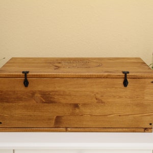 May include: A wooden box with a hinged lid and two black metal clasps. The box is stained a dark brown color and has a small inscription on the top.