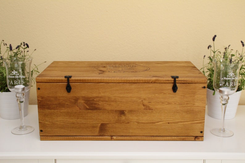May include: A wooden box with a hinged lid and two black metal clasps. The box is stained a dark brown color and has a small inscription on the top.